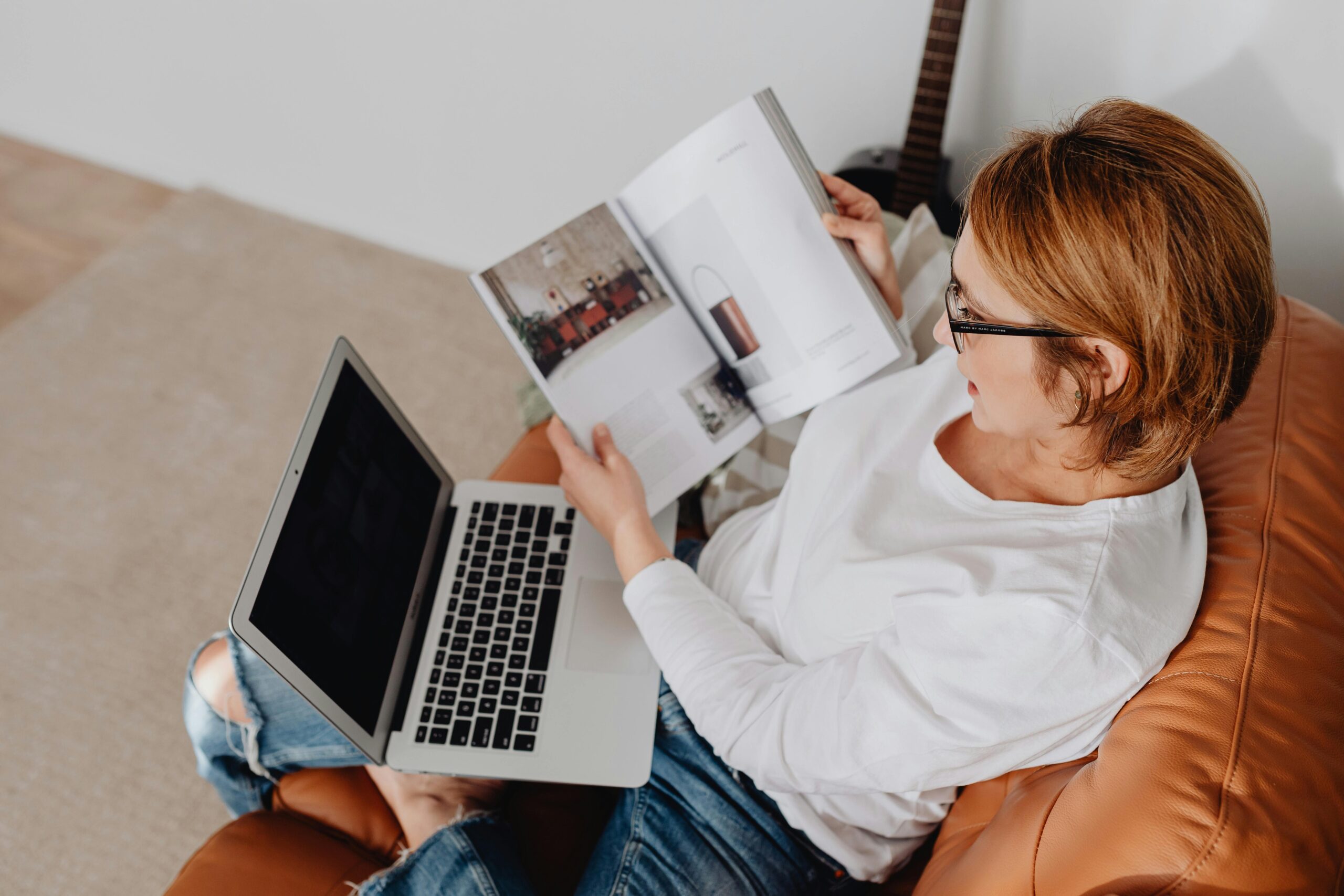 A woman sitting on a couch reading a magazine and using a laptop in a cozy living room.